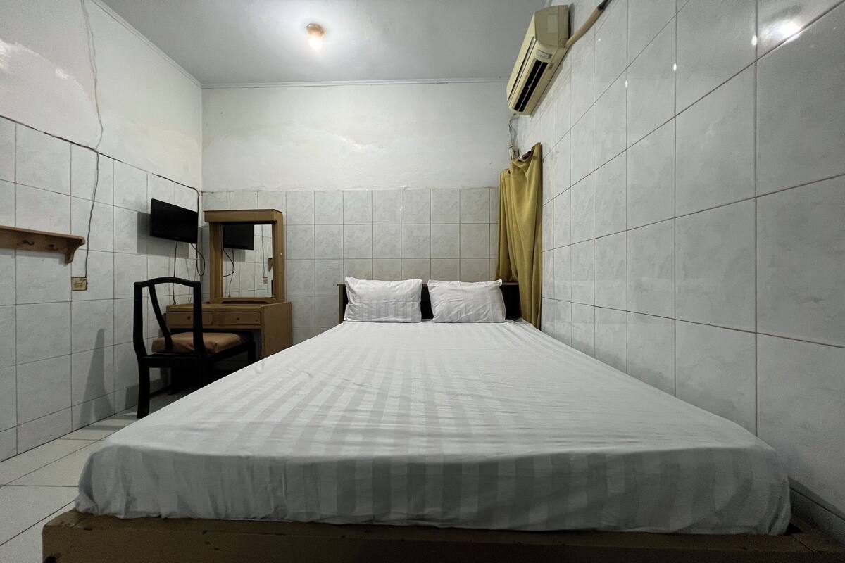A small, clean room with white tiled walls, a bed with white bedding, a wooden nightstand, a chair, a mirror, and an air conditioning unit.
