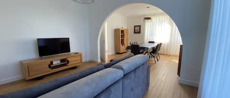 A modern living room with a TV on a wooden console, a grey sofa, and a dining area with a table and chairs.