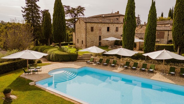 A pool area with lounge chairs and umbrellas, surrounded by stone buildings and tall trees. - Villa Camelia by Elite Villas (Colle di Val d'Elsa)