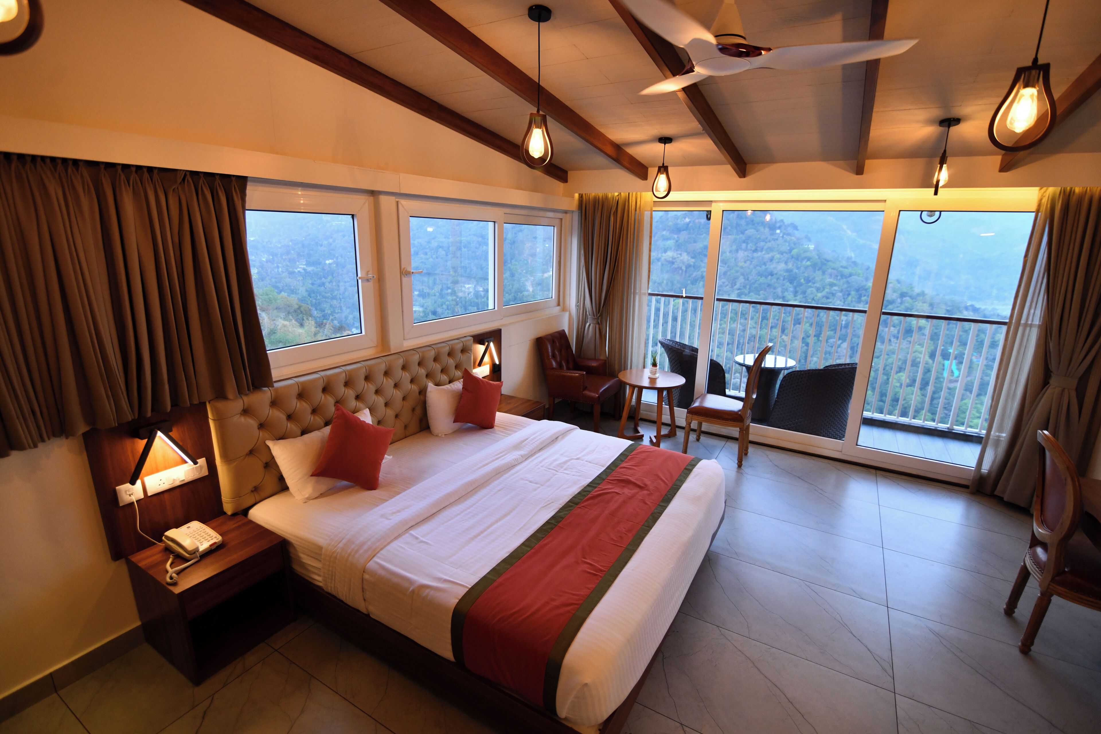 A hotel room with a large bed, a nightstand with a phone, a chair, a small table, and a balcony with a view.