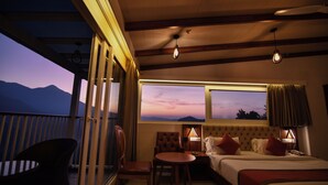 A hotel room with a bed, a table, two chairs, a window with a view of mountains, and a balcony. - British County Resort (Devikolam)