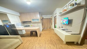 Eine kompakte Küche mit weißen Schränken, einem Kühlschrank, einer Mikrowelle und einer kleinen Arbeitsfläche. Der Boden ist aus Holz und an der Wand ist ein Fernseher befestigt.