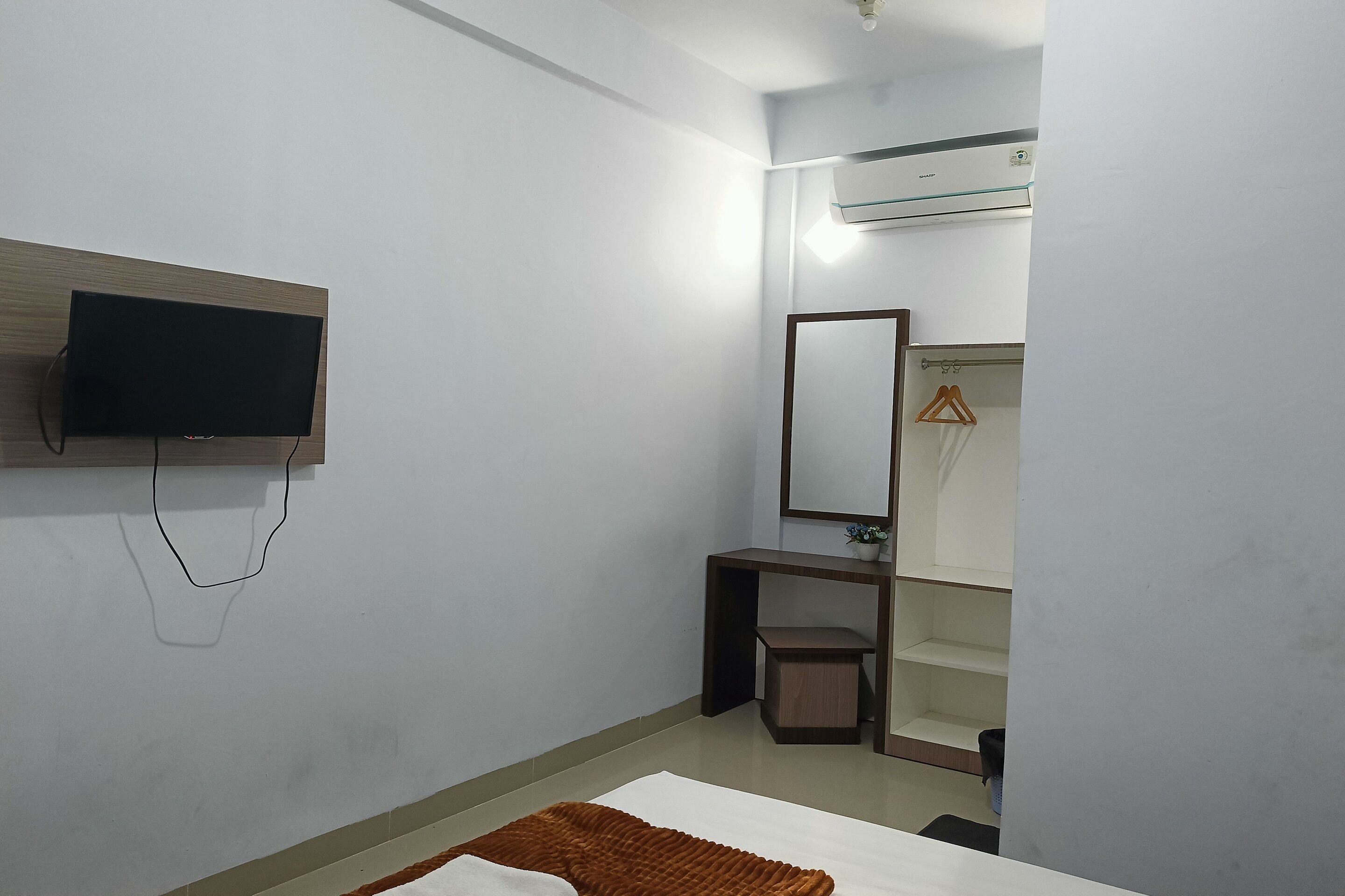 Quarto de hotel com cama, televisão na parede, pequena escrivaninha, espelho e ar condicionado.