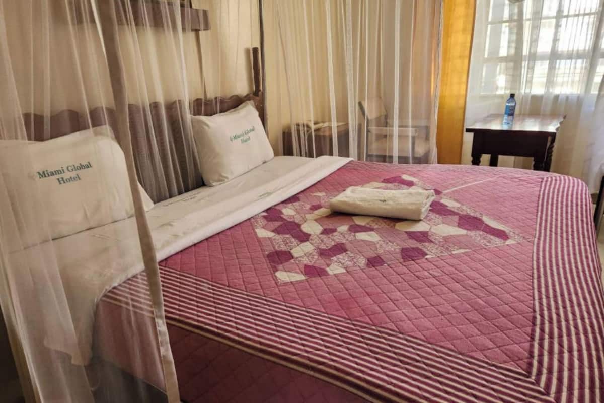 A hotel room with a bed covered in a red quilt, a white pillow with "Miami Global Hotel" text, and a mosquito net.