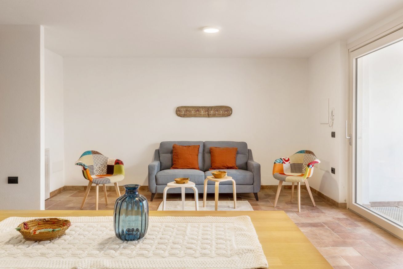 A living room with a grey sofa, two small white tables, colorful chairs, a blue vase, and a white knitted rug.