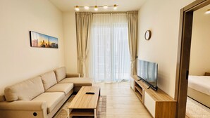 Een moderne woonkamer met een bank, een salontafel, een tv op een houten dressoir en een groot raam met dunne gordijnen.