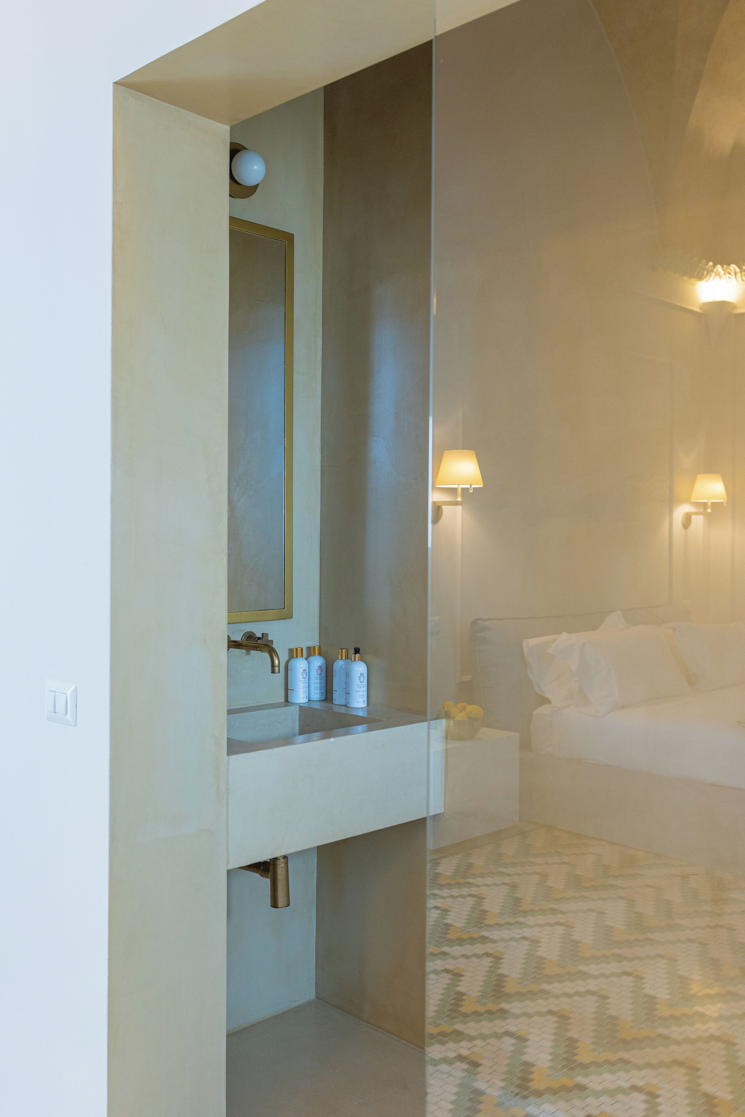 A bathroom with a mirror, sink, and toiletries. There is a bed with a white comforter and pillows in the adjacent room.