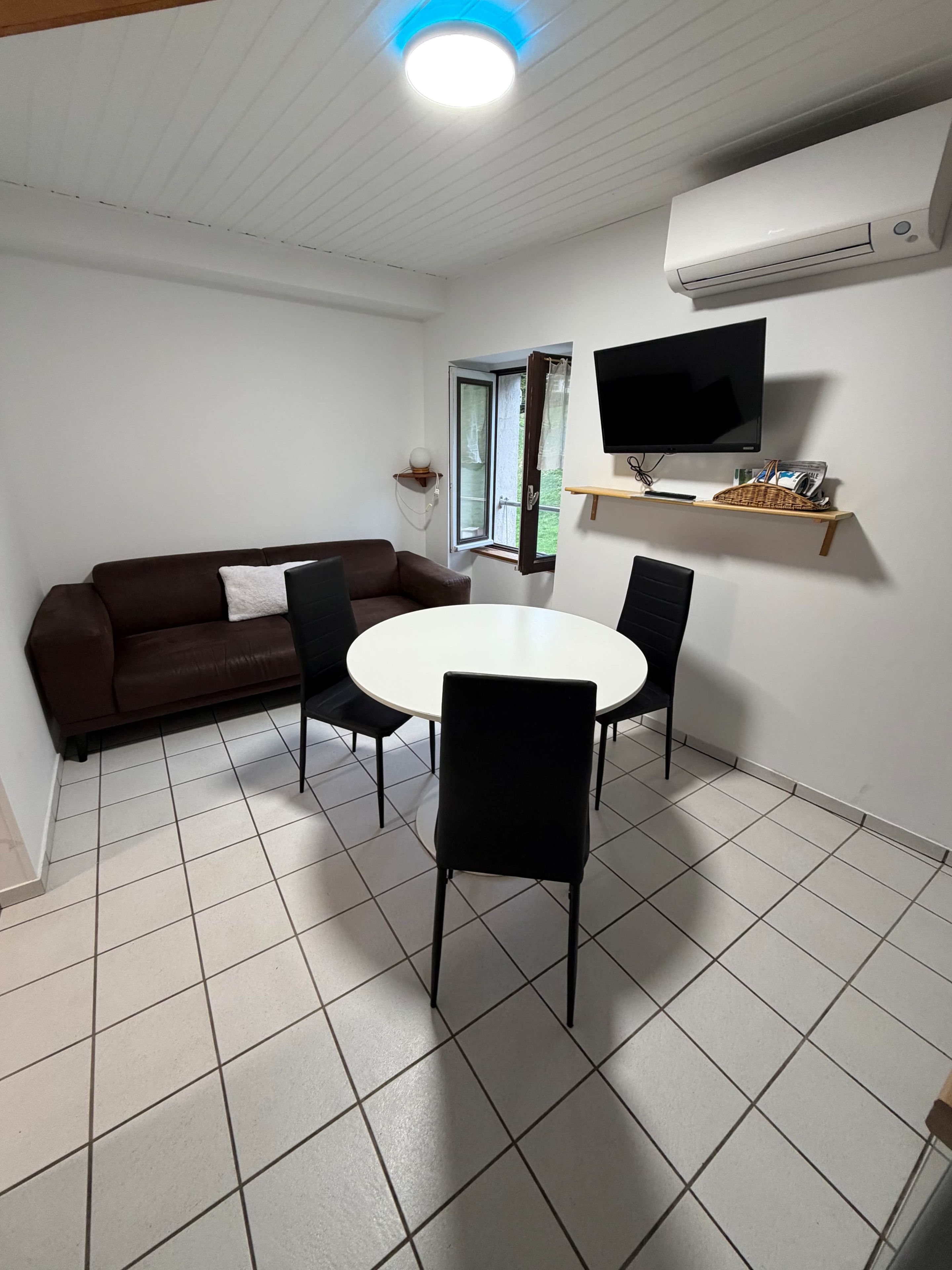 A room with a brown sofa, a white round table with four chairs, a television on a wall-mounted shelf, and an air conditioning unit.