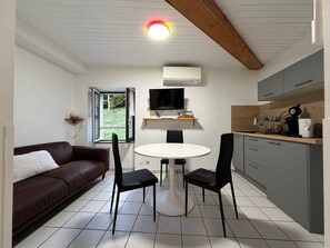 Een knus woongedeelte met een kitchenette, een eethoek met een ronde tafel en stoelen, een bruine bank en een aan de muur bevestigde televisie.