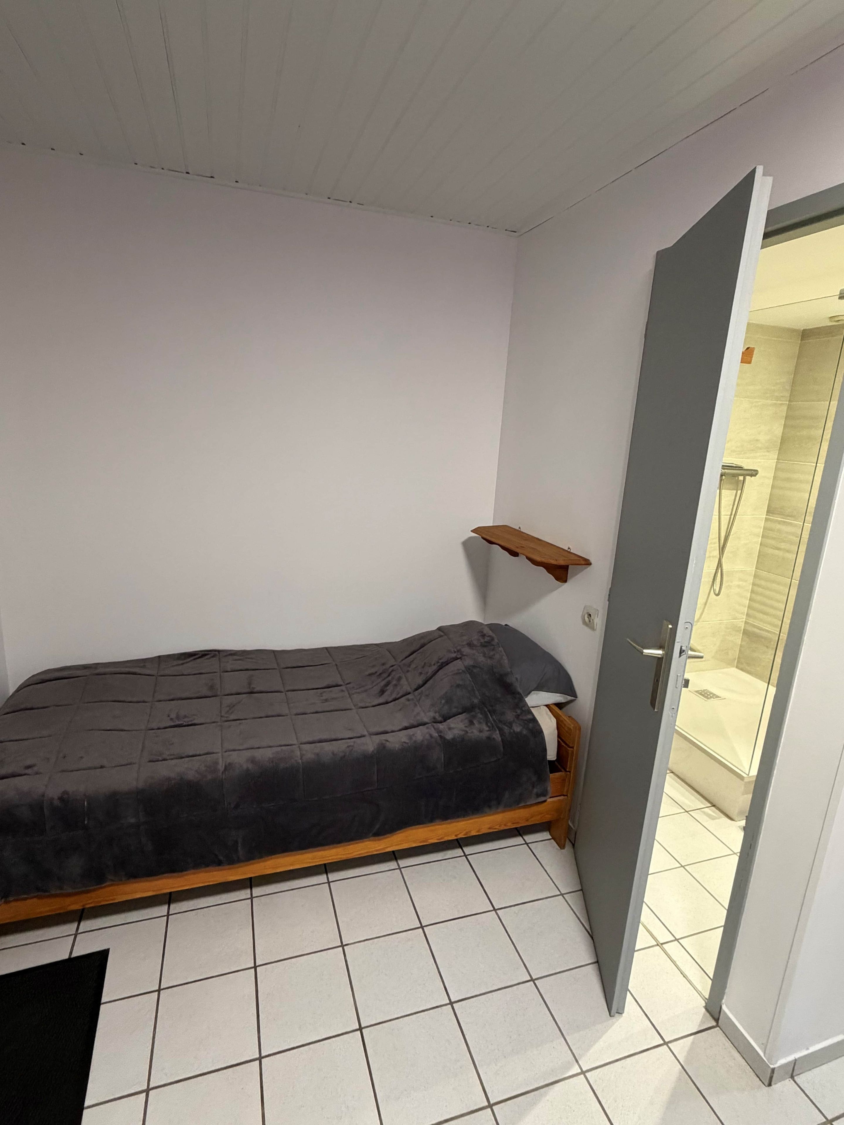 Een kleine kamer met een enkel bed, een houten frame, een grijze matras en een vloer van witte tegels.