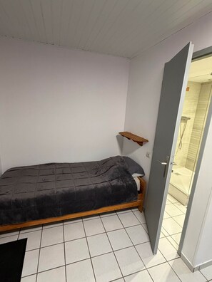 A small, single-bed room with a wooden frame, a grey mattress, and a white tiled floor.