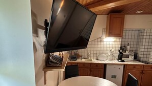 A compact kitchen with a round table, black chairs, a TV mounted on the wall, and basic appliances. - Aurore - Appartement 3 Personnes (Saint-Martin-d'Uriage)