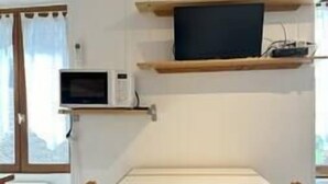 A room with a wooden table and chairs, a flat-screen TV on a shelf, a microwave, and an air conditioner. - Alhambra - Studio Cosy (Saint-Martin-d'Uriage)