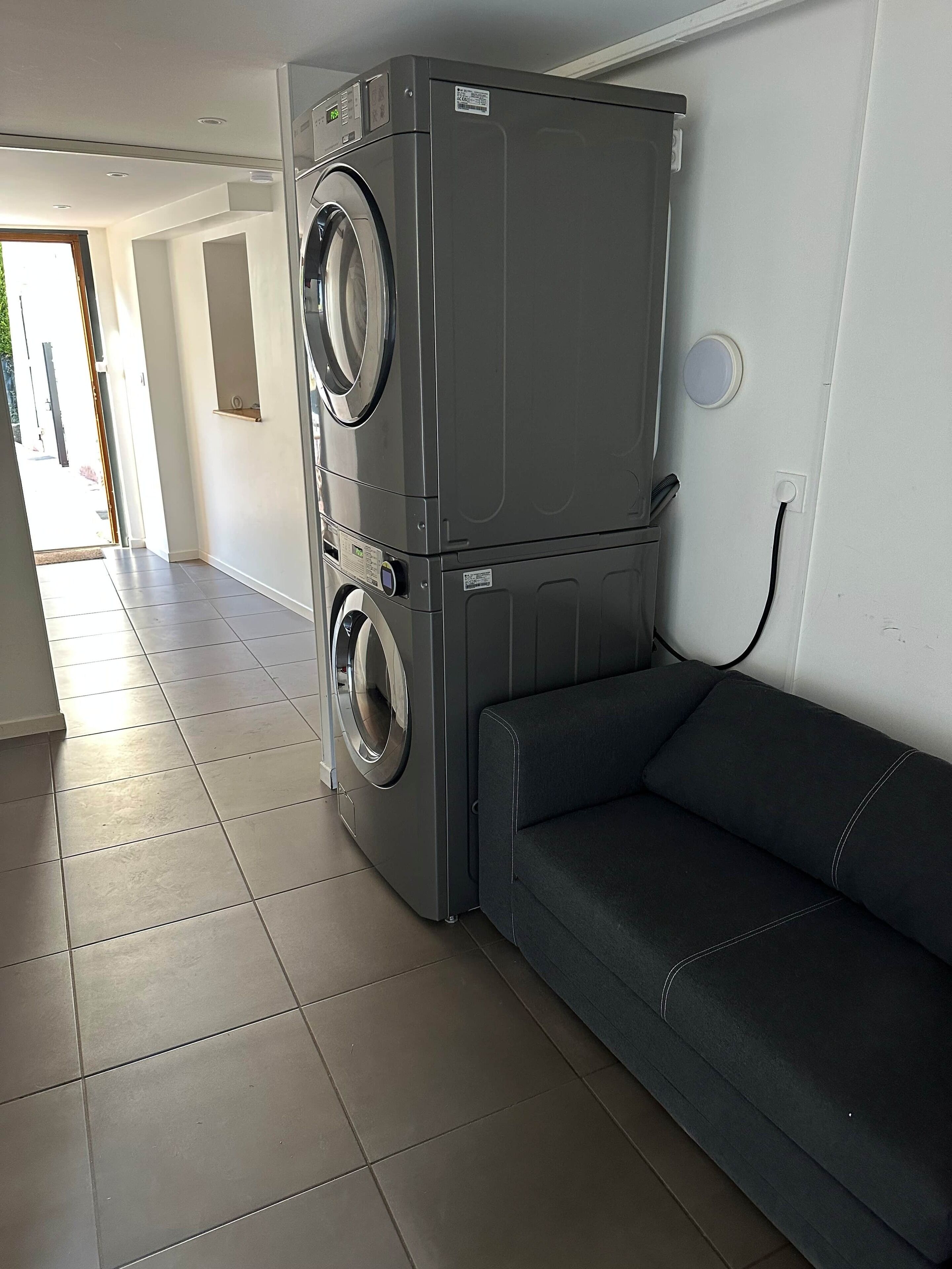 A compact laundry room with a stacked washer and dryer, a dark-colored sofa, and a tiled floor.