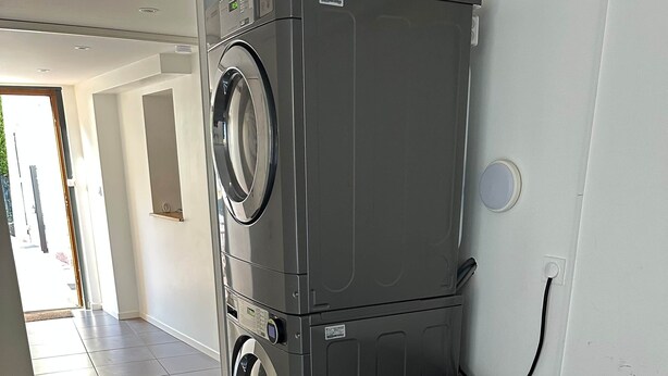 Une petite buanderie équipée d’un lave-linge et d’un sèche-linge superposés, d’un canapé de couleur foncée et d’un sol carrelé.