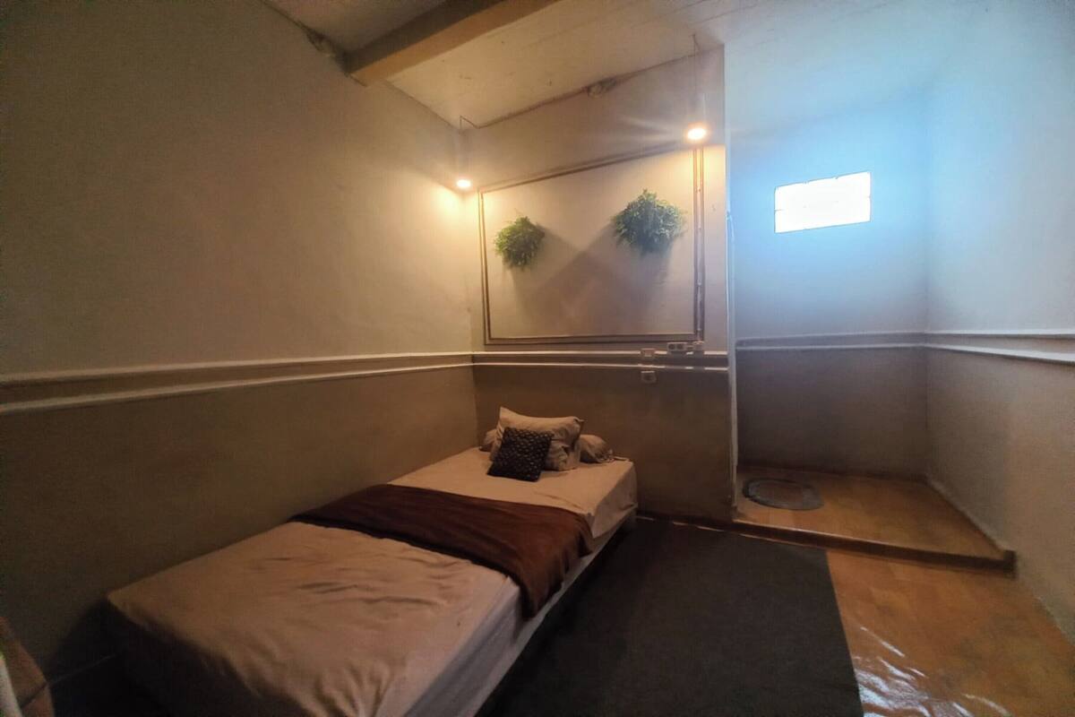 Ein kleiner, schummrig beleuchteter Raum mit einem Einzelbett, einem kleinen Fenster und einem Spiegel, der eine Pflanze reflektiert.