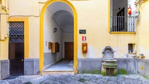 Una estrecha calle con un edificio amarillo, una puerta negra y una fuente de piedra.