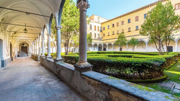 Un pasaje cubierto con arcos, un patio con setos podados y un edificio con múltiples ventanas.