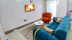 Ein Wohnzimmer mit einem blauen Sofa, einem orangefarbenen Sessel, einem an der Wand montierten Fernseher und einem kleinen weißen Tisch.
