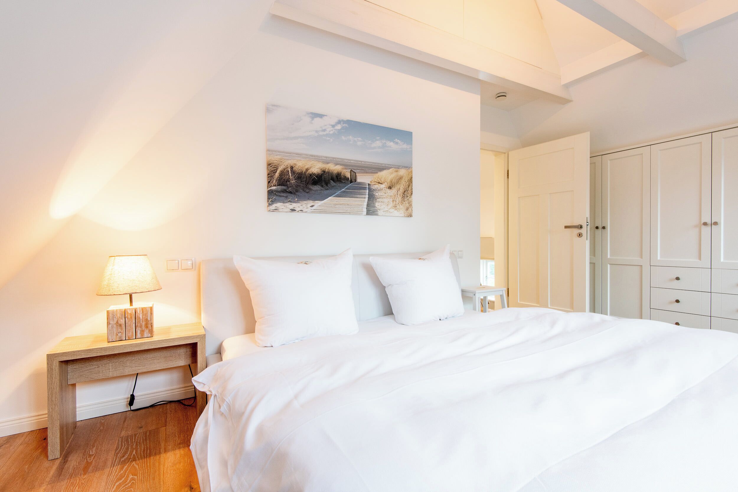 A bedroom with a large bed, white walls, a painting of a beach, and a wardrobe.