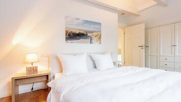 A bedroom with a large bed, white walls, a painting of a beach, and a wardrobe.