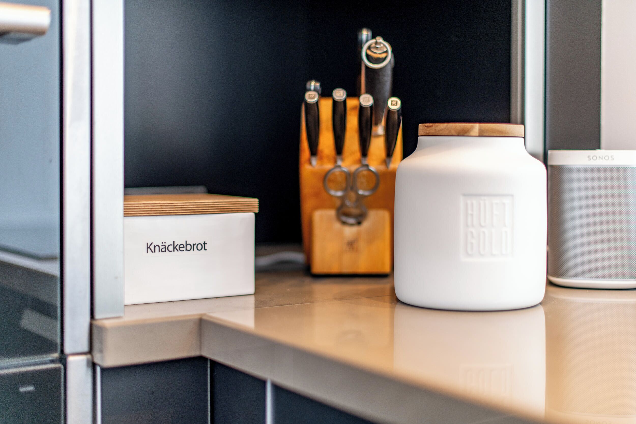 A kitchen counter with a white storage box labeled "Knäckebrot," a wooden knife block, a white container with "Ruft Gold" branding, and a SONOS speaker.
