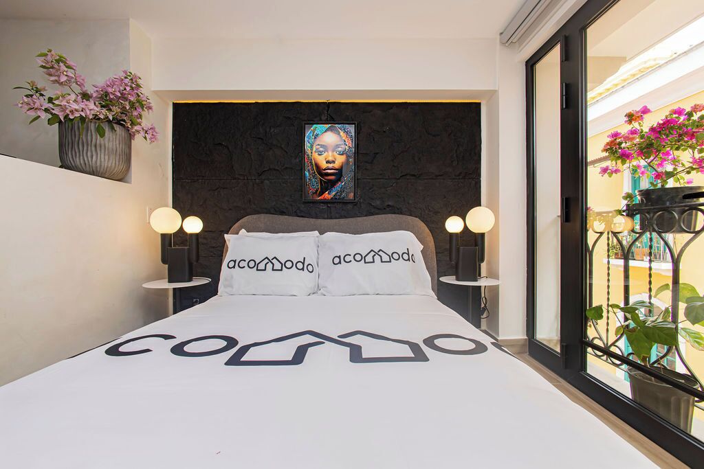 A modern bedroom with a bed featuring a geometric design, two bedside lamps, a dark headboard, and a framed artwork above the bed.