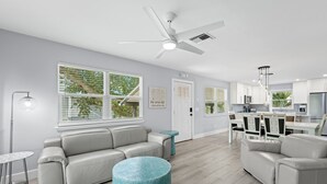 Una sala de estar moderna con un sofá gris, un otomano azul redondo y un comedor con una mesa y sillas blancas.