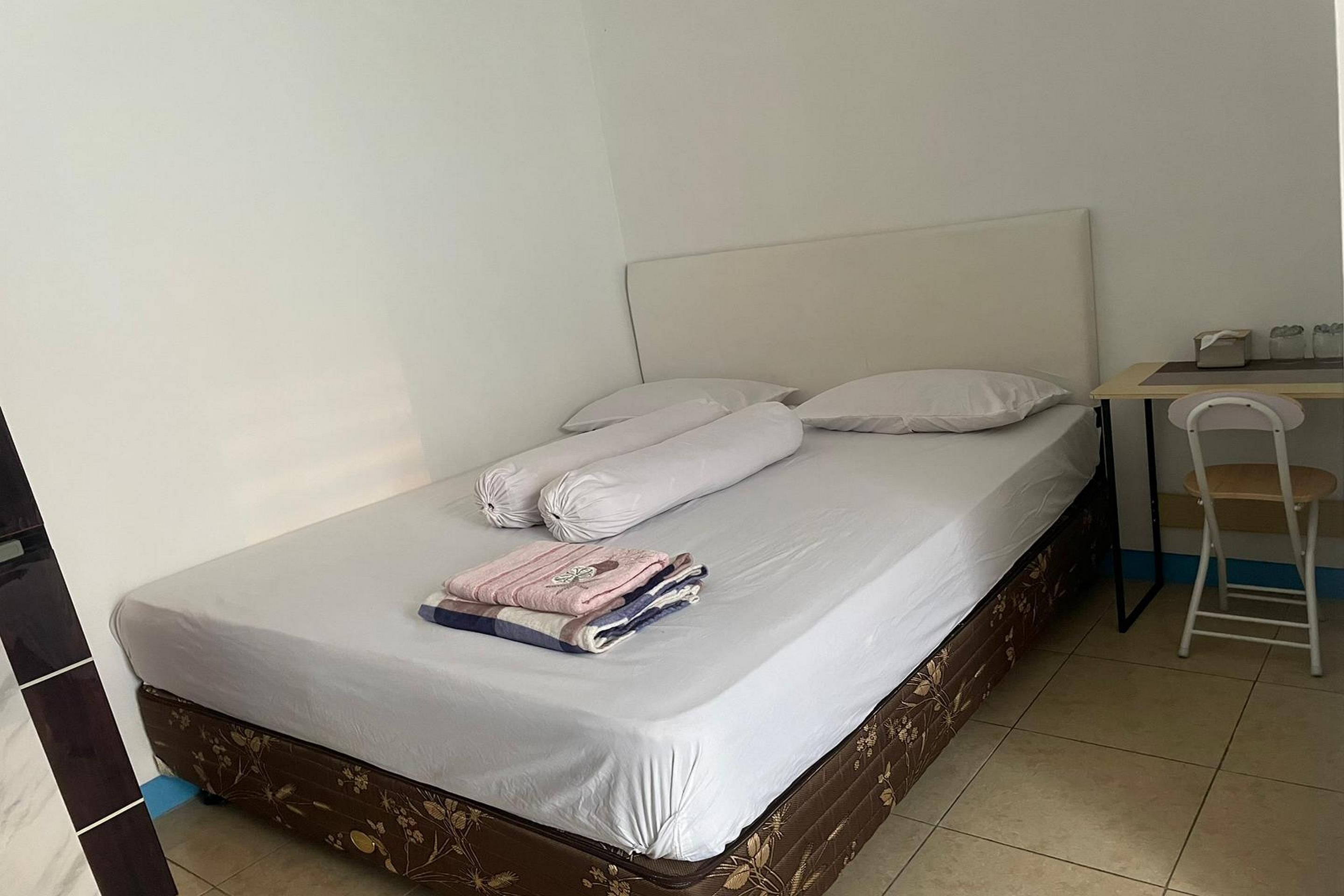 A neatly made bed with pillows and a folded blanket, a small table with a chair, and a tiled floor.