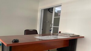Una scrivania in legno con un telefono e documenti, una sveglia al muro e una porta con un cartello.