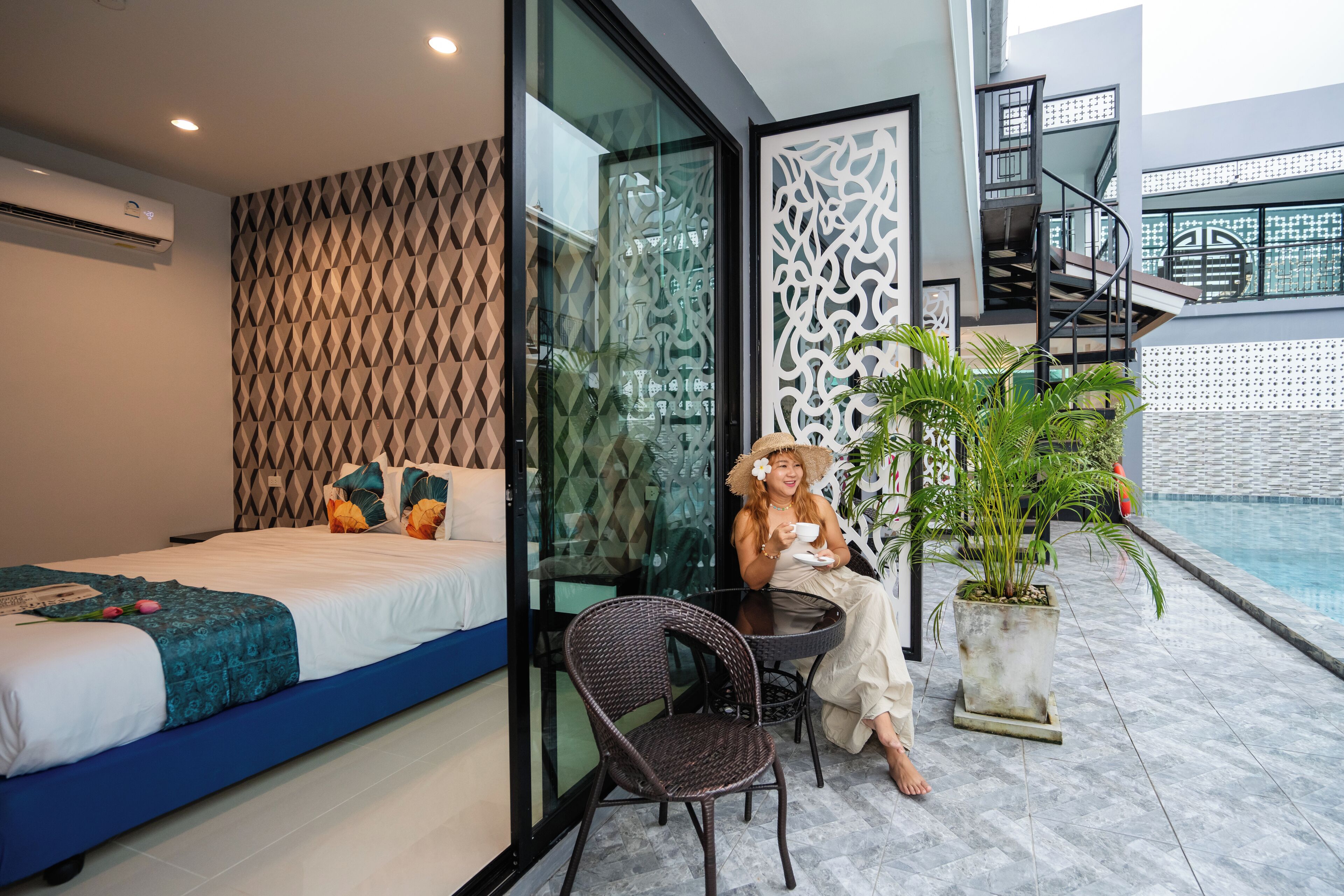 A modern hotel room with a bed, a glass partition, a dining area with a chair, and a potted plant.