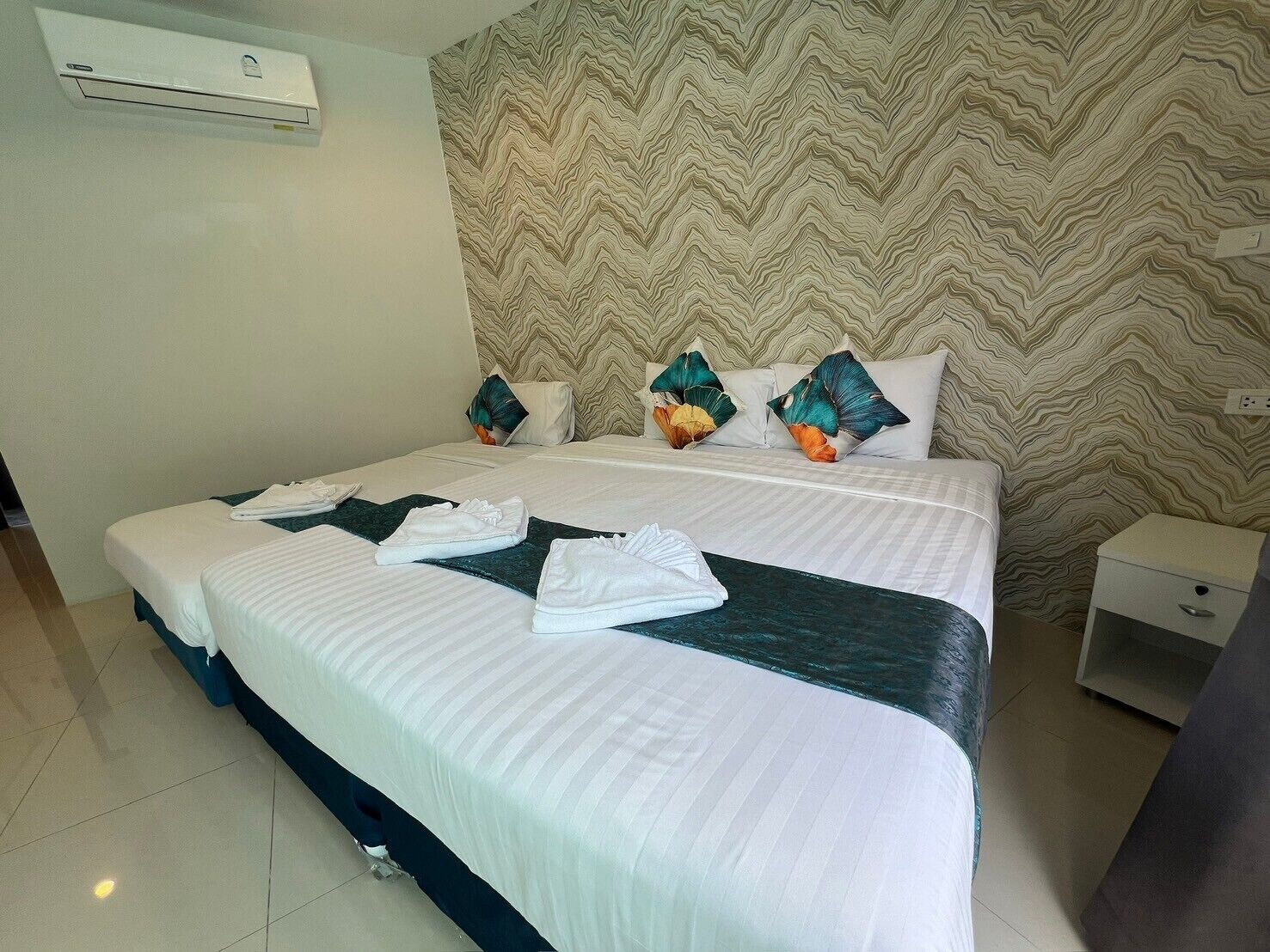 A hotel room with a large bed, a nightstand, a wall-mounted air conditioner, and a patterned wallpaper.
