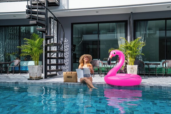 A woman sitting by a pool with a pink flamingo float, surrounded by potted plants and outdoor furniture.