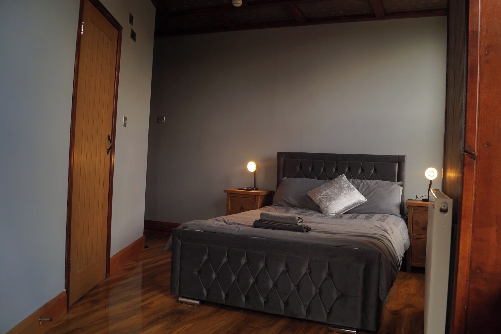Un dormitorio con una cama tapizada oscura, dos mesitas de noche con lámparas, una puerta de madera y suelo de madera.
