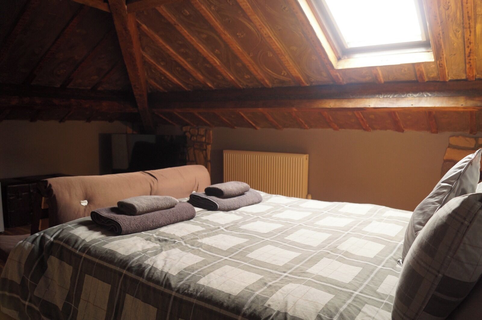 A bedroom with a sloped ceiling, a bed with a checkered bedspread, a radiator, and a window with a view of the sky.