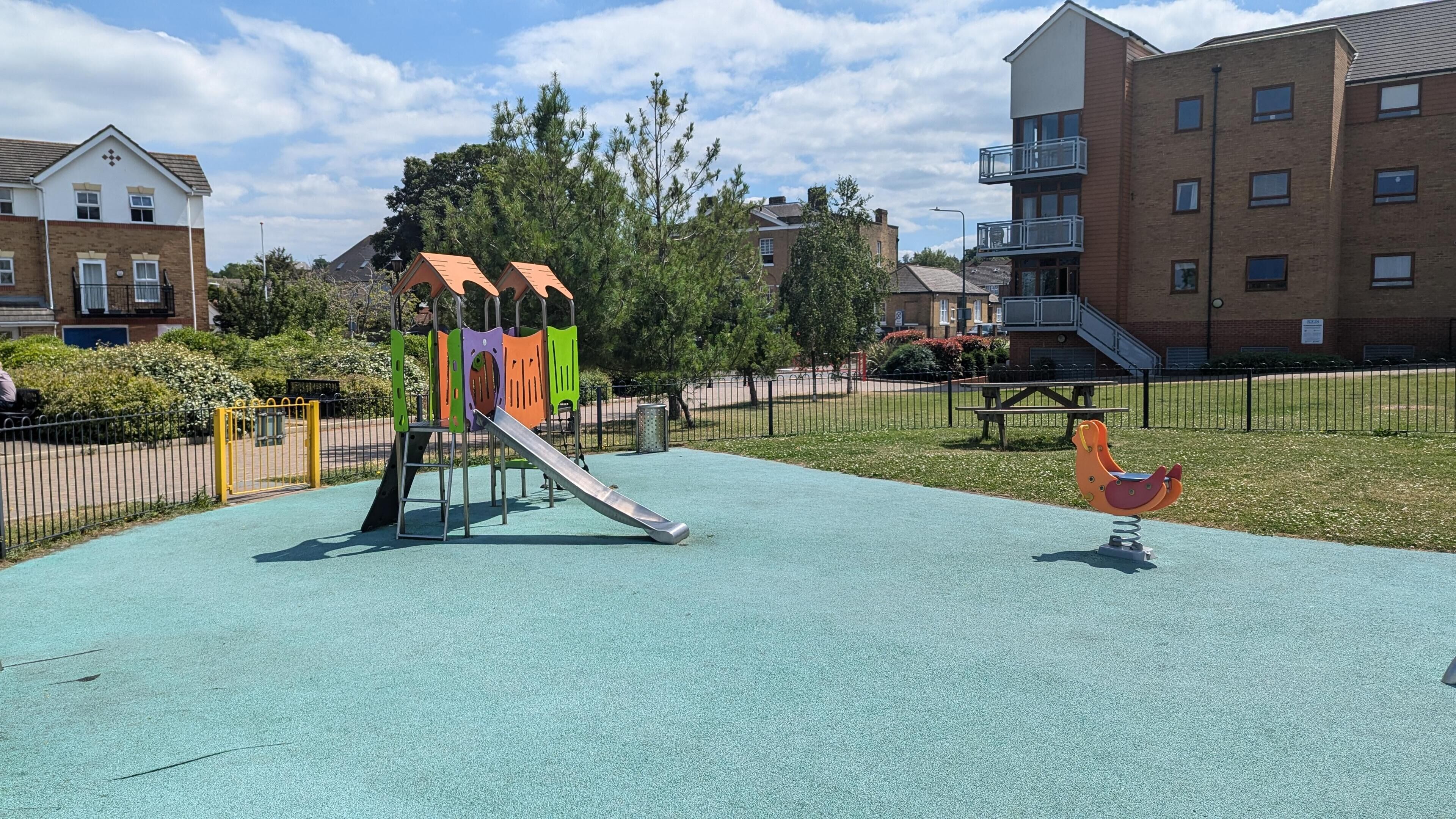 Un terrain de jeux avec un toboggan et des balançoires, entouré d’une clôture et de verdure.