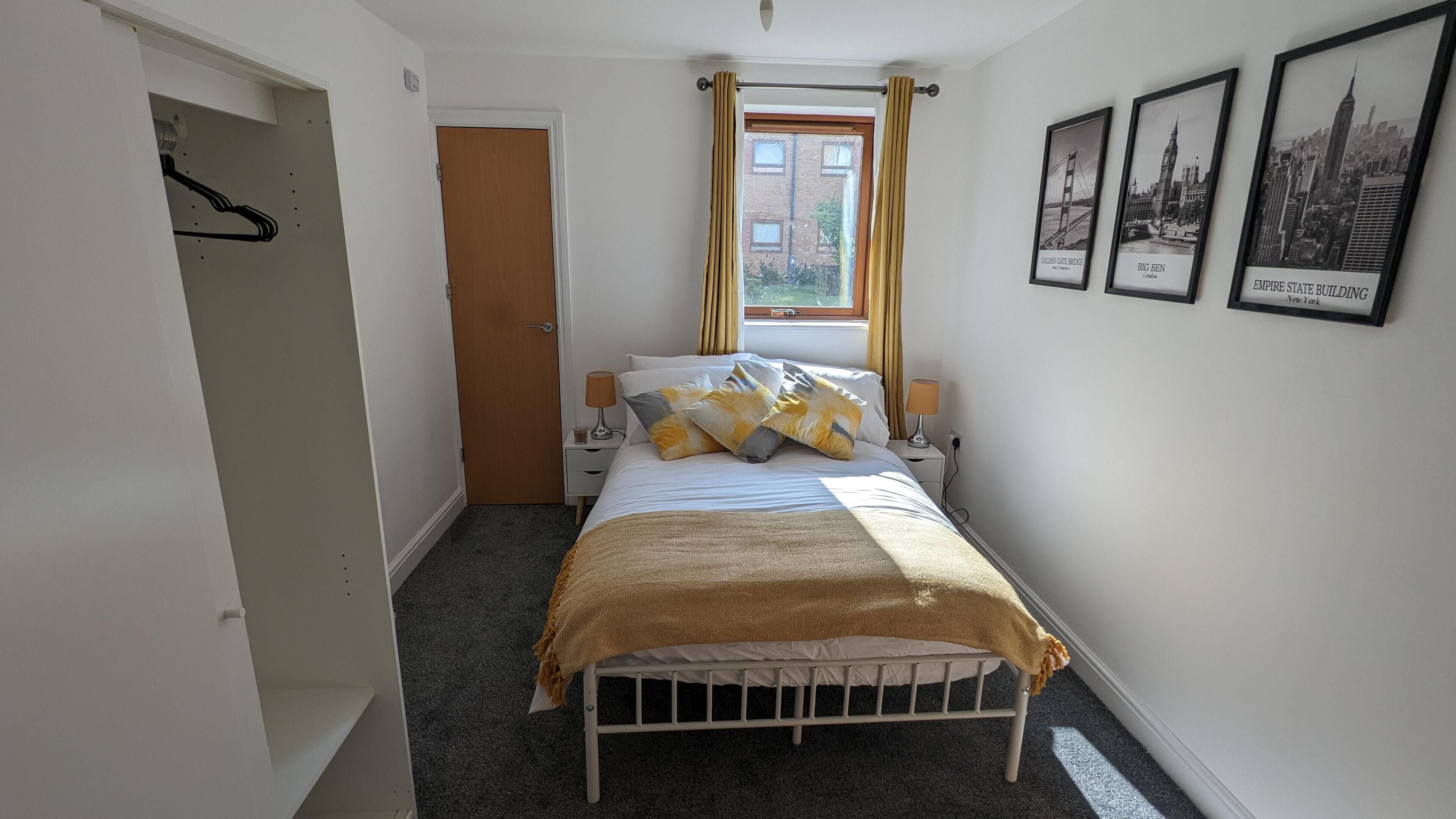 A bedroom with a bed, a window with curtains, a door, and framed pictures on the wall.