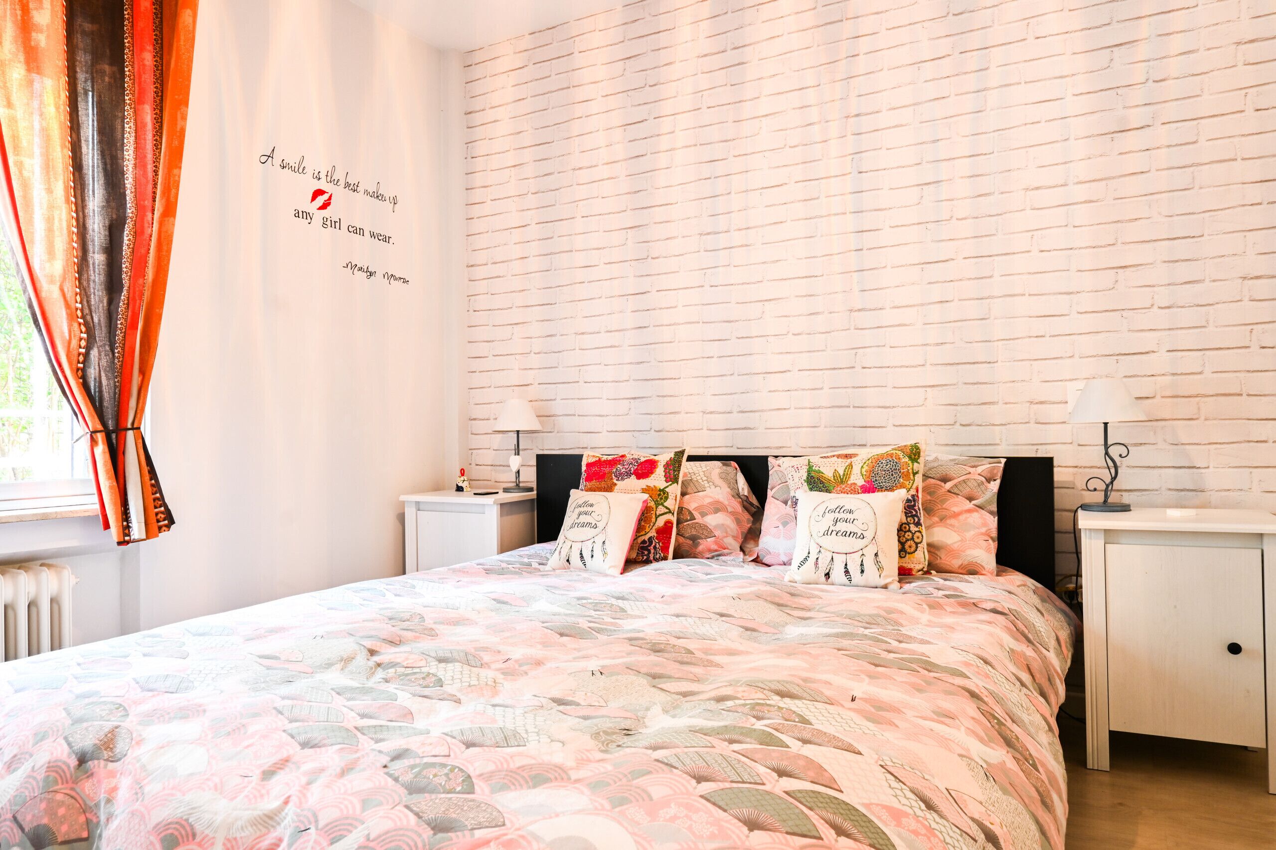 Un dormitorio con una pared de ladrillo visto, una cama con colcha estampada, dos mesitas de noche y una ventana con cortinas.