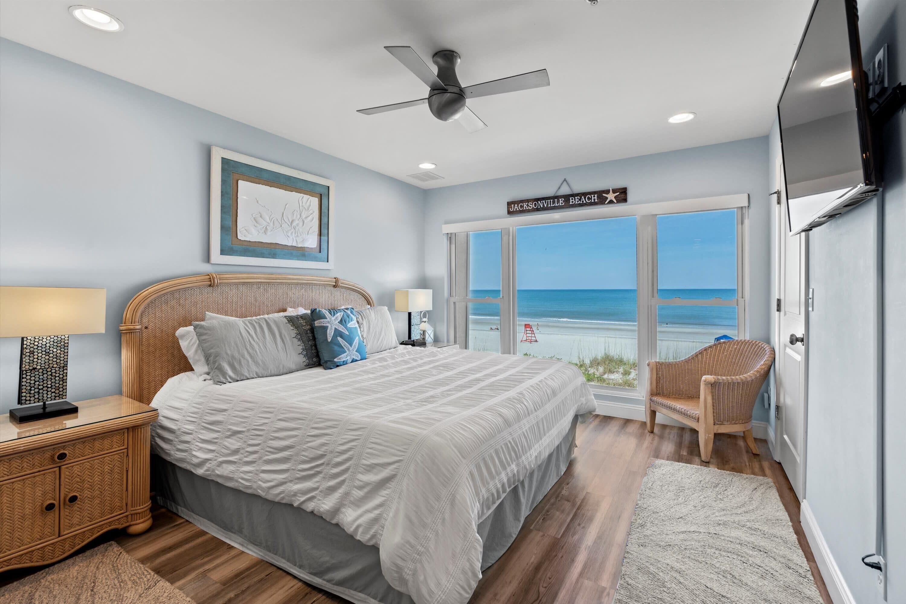 Un dormitorio con una cama grande, un ventilador de techo, una mesita de noche con lámpara, un sillón de mimbre y vista a la playa.