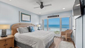 Una camera da letto con un letto grande, una ventola a soffitto, un comodino con una lampada, una sedia in vimini e vista sulla spiaggia.