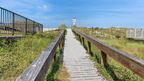 Una passerella in legno conduce a una spiaggia, con una recinzione metallica da un lato e una zona erbosa dall'altro.