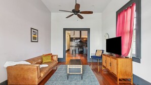 A living room with a brown leather sofa, a wooden TV stand, a ceiling fan, and a view into the kitchen.