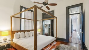 A four-poster bed with a ceiling fan, a glass table with a lamp, and a view into the hallway.