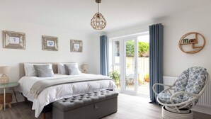 Ein Schlafzimmer mit einem Bett, Nachttischen, einem grauen Pouf, einem Korbstuhl und einem großen Fenster mit Vorhängen.