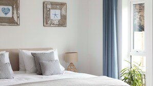 Ein Schlafzimmer mit einem großen Bett, einem grauen Hocker, einer hängenden Lampenschirm aus Weide und einem Fenster mit Vorhängen.