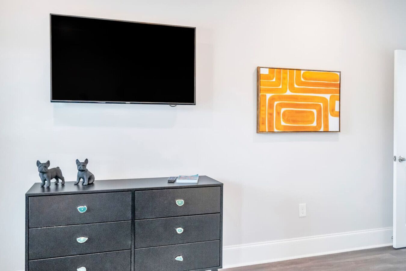 A modern room with a flat-screen TV mounted on the wall, a dark-colored dresser with decorative items, and a geometric abstract painting.
