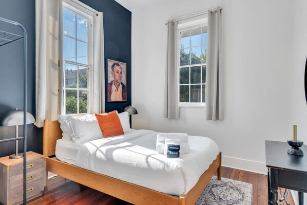 A bedroom with a wooden bed, white bedding, a bedside table with a lamp, and a small desk with a lamp.