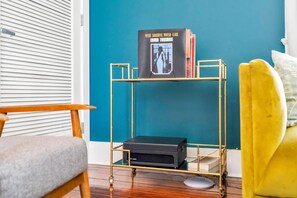 A room with a teal wall, a gold-framed bookcase with a vinyl record, a black stereo system, a wooden chair, and a yellow armchair. - Hip & Stylish Magazine St. Apt | Balcony & Parking (New Orleans)