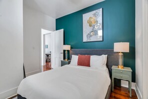 A bedroom with a large bed, two bedside tables with lamps, a teal accent wall, and a framed artwork above the bed.