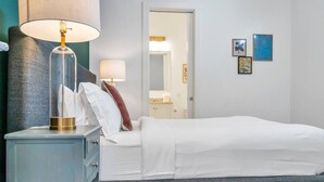 A neatly made bed with white linens, a bedside lamp, and a small table with a lamp. There are two framed artworks on the wall.